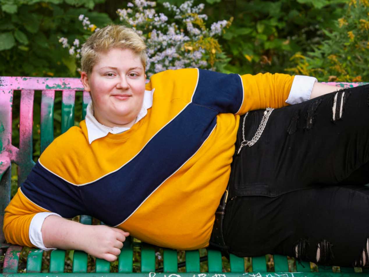 A photo of a young, large, white woman with a bleached blond crew cut looking at the camera wearing a yellow sweater with a big navy blue horizontal stripe over a white collared shirt laying on one arm on a park bench.