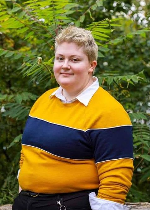 A photo of a young, large, white woman with a bleached blond crew cut looking at the camera wearing a yellow sweater with a big navy blue horizontal stripe over a white collared shirt standing in front of a green bush.